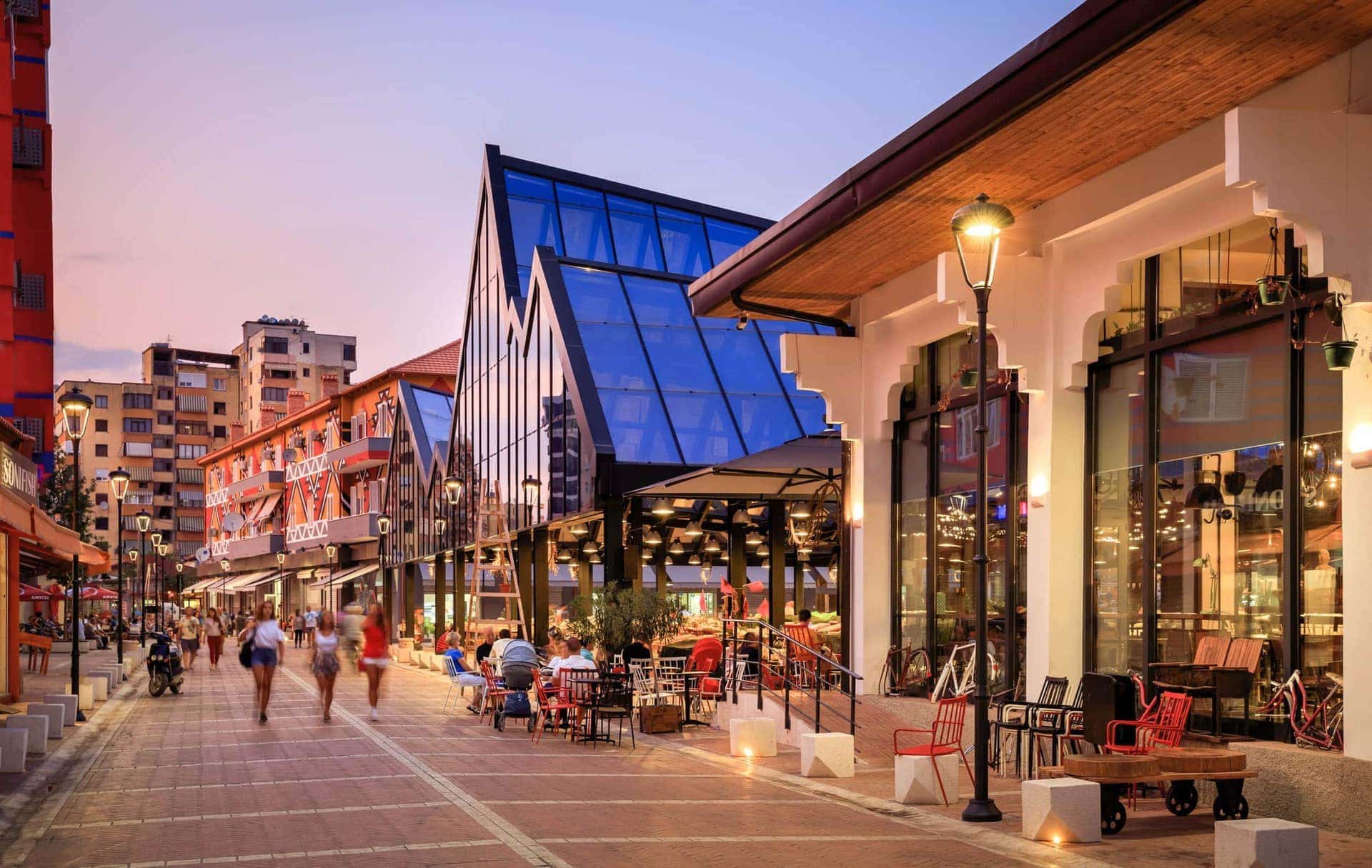 Vibrant evening at Tirana's Bazaar with people dining under red awnings, next to modern glass buildings reflecting the sunset sky.